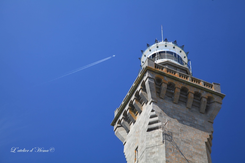 Vue en contreplongée du phare d'Eckmühl. Un avion passe dans le ciel bleu à l'arrière plan