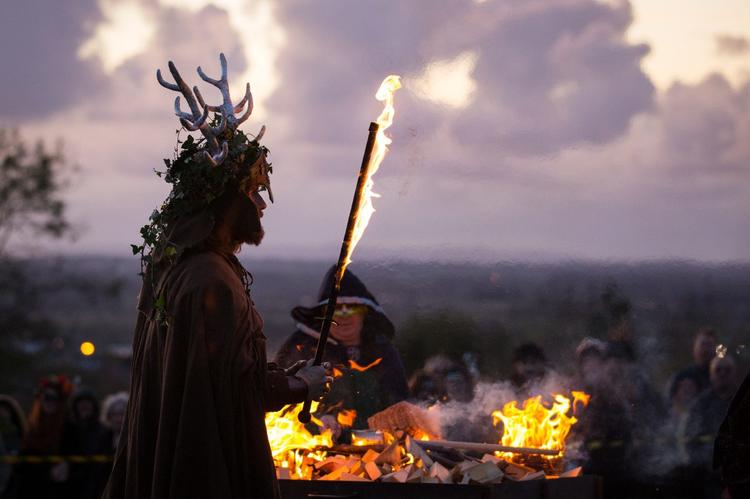 Reconstitution de la fête celte Samain à Glastonbury dans le Royaume-Uni. Au premier plan, un homme costumé en druide tient un bâton en feu à la nuit tombée devant un feu de joie entouré d'autres acteurs.